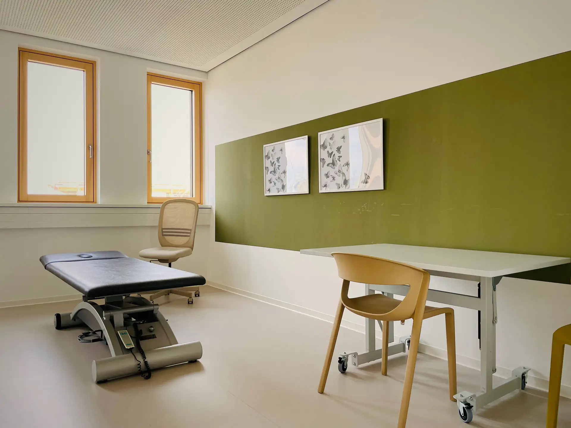 Heller Ergotherapieraum im Therapie Zentrum Augsburg, ausgestattet mit einer Therapieliege, einem Schreibtisch und Stühlen, bietet eine ruhige Atmosphäre für therapeutische Sitzungen.