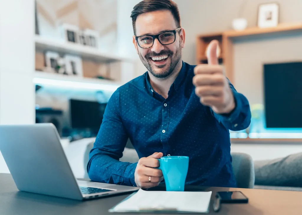 Fröhlicher Mann mit Brille und Bart zeigt einen Daumen hoch, während er eine blaue Tasse hält, als Symbol für Zufriedenheit und Erfolg im Rahmen der betrieblichen Gesundheitsförderung.