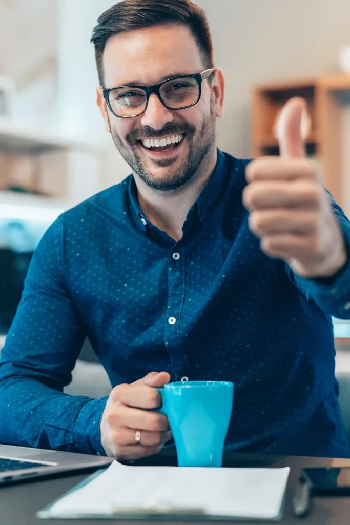 Fröhlicher Mann mit Brille und Bart zeigt einen Daumen hoch, während er eine blaue Tasse hält, als Symbol für Zufriedenheit und Erfolg im Rahmen der betrieblichen Gesundheitsförderung.