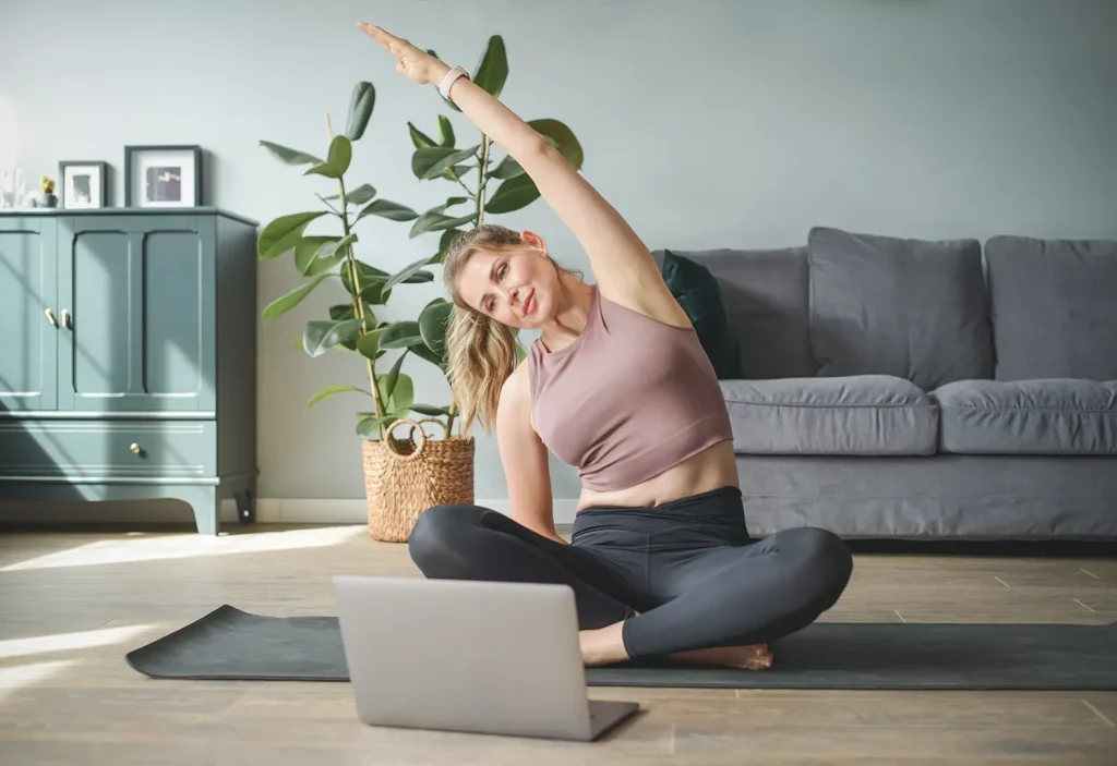 Junge Frau, die eine seitliche Dehnübung macht und dabei auf einem Laptop Yoga-Übungen folgt, symbolisiert die Integration von körperlicher Fitness und Wohlbefinden in die betriebliche Gesundheitsförderung.