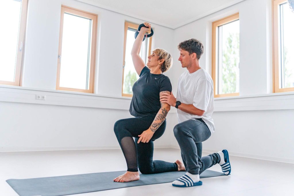 Trainer begleitet Patientin bei funktionellem Training mit Kettlebell auf dem Gesundheitscampus, individuelle Bewegungstherapie in heller Trainingsumgebung