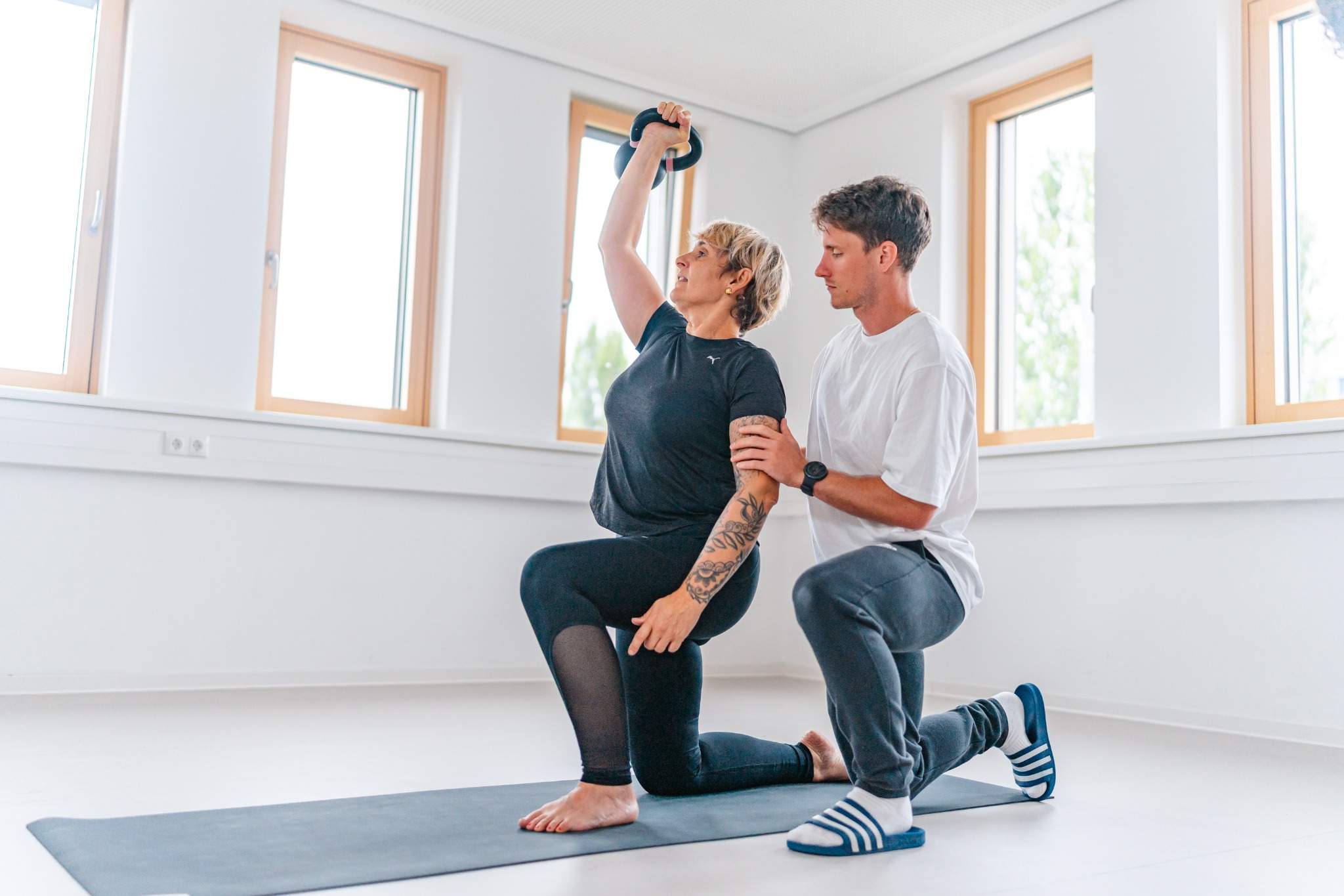 Trainer begleitet Patientin bei funktionellem Training mit Kettlebell auf dem Gesundheitscampus, individuelle Bewegungstherapie in heller Trainingsumgebung